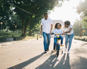 An image of a family teaching their child how to ride a bike, symbolic of them teaching them all the steps of growing up, including helping them with financial planning and saving by giving her pocket money.